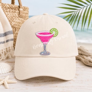 May include: A beige baseball cap featuring a beaded patch of a pink margarita glass with a white rim and a green lime slice garnish. The glass stem and base are covered in silver glitter. The text Brazenly Envied is printed across the center.