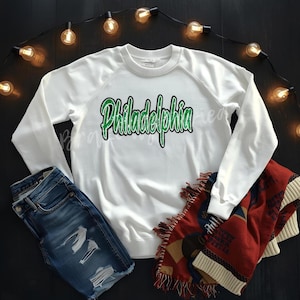 May include: A white sweatshirt with the word "Philadelphia" in green script. The sweatshirt is paired with blue denim shorts and a red and beige patterned blanket. String lights are in the background.