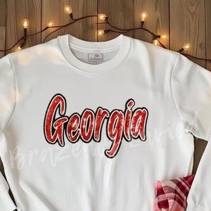 May include: A white sweatshirt with the word "Georgia" in red, outlined in black, across the chest. The long-sleeved top is made of a soft material and is suitable for casual wear. The background is a wooden surface with string lights.