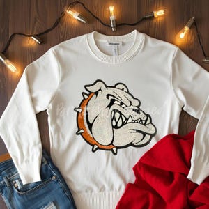 May include: White sweatshirt with a sequined bulldog graphic. The bulldog is outlined in orange and has a fierce expression. The sweatshirt is paired with blue jeans and a red hoodie. Decorative string lights are in the background.