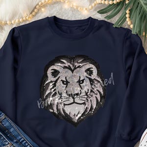 May include: Navy blue sweatshirt featuring a sequined lion's head design. The lion's mane is black, and the face is silver. The text "Paired" is written in cursive on the right side of the lion's head.