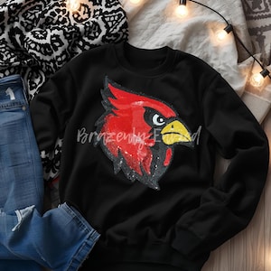 May include: Black sweatshirt featuring a large, sequined cardinal bird design in red, black, and yellow. The text "Brazenly Edward" is printed on the sweatshirt. The sweatshirt is displayed on a neutral background with decorative lights.
