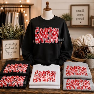 May include: A black crewneck sweatshirt displayed on a mannequin, featuring the word GAMEDAY in red with white polka dots. Below, two folded black sweatshirts, one white sweatshirt, and two gray sweatshirts all feature the same red and white graphic.