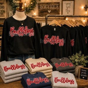 May include: A collection of black, gray, beige, and navy blue crewneck sweatshirts featuring the word Bulldogs in red glitter script lettering. The sweatshirts are displayed on a mannequin and in neatly folded stacks on a wooden surface.