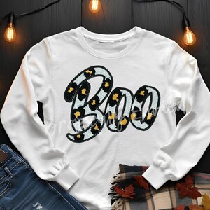 May include: White sweatshirt with the word "Boo" in a leopard print design. The letters are outlined in a light blue color. The sweatshirt is paired with blue jeans and a fall-themed scarf.