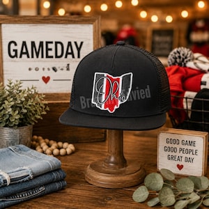 May include: A black trucker hat featuring a patch shaped like the state of Ohio. The patch displays a red, white, and black brushstroke design with the word Ohio written in a white script font across the center of the state shape.