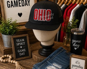 May include: A black trucker-style baseball cap featuring the word OHIO embroidered in bold red letters with a white outline on the front panel. The hat has a structured crown, a flat brim, and a mesh back panel, displayed on a neutral mannequin head.
