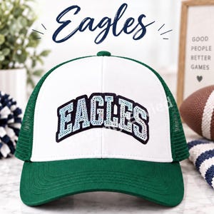 May include: A white and green trucker hat featuring the word EAGLES in a dark blue and light blue sequined font across the front panel. The cap has a solid white front, a green brim, and green mesh side and back panels with a button on top.