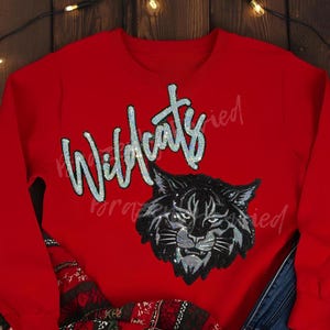 May include: A red sweatshirt featuring the word "Wildcats" in a sparkling, cursive font. Below the text is a black and silver sequined image of a wildcat's face. The sweatshirt is a solid red color.