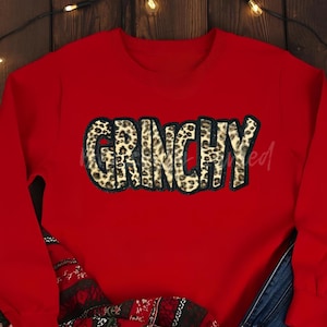 May include: A red sweatshirt with the word "GRINCHY" in a leopard print design across the chest. The sweatshirt is a solid red color, and the text is outlined in black.
