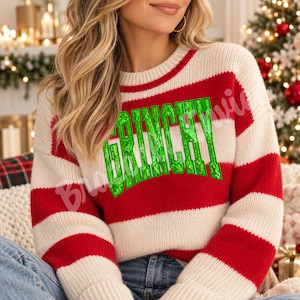 May include: A long-sleeve knit sweater featuring horizontal red and cream stripes. The center of the chest displays the word GRINCHY in large, bright green, glittery, block-style letters with a distinct textured appearance.