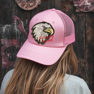 May include: Pink trucker hat with a mesh back and a sequined eagle patch. The eagle has silver, black, and yellow details. The text "Brodly Married" is in red below the eagle.