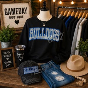 May include: A black long-sleeve sweatshirt with the word BULLDOGS in blue and silver sequins, a black baseball cap with the word BULLDOGS in blue, blue distressed denim jeans, a tan fedora, and a black tumbler with the text game day.
