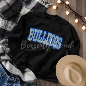 May include: Black sweatshirt with the word "BULLDOGS" in a blue sequined design. The word "Brazenly" is written in cursive below the word "BULLDOGS". A tan hat and a plaid blanket are also in the image.