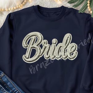 May include: Navy blue sweatshirt with the word "Bride" in a cursive, sequined design. The text is outlined in black and has a silver-white color. The sweatshirt is a crew neck style and is paired with blue jeans and white sneakers.