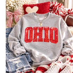 May include: A heather gray crewneck sweatshirt featuring the word OHIO in large, red, glittery varsity-style letters across the chest. The garment has long sleeves and a ribbed neckline, paired with a gold-toned pendant necklace.