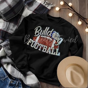 May include: Black sweatshirt with the words "Bulldogs Football" in a sequined design. The design includes a football and a bulldog graphic. A tan hat and plaid blanket are in the background.