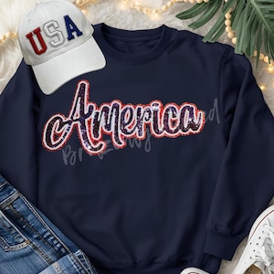 May include: Navy blue sweatshirt with the word "America" in sequined script. A white baseball cap with "USA" in red and blue lettering sits above the sweatshirt. The image also includes blue jeans and white sneakers.