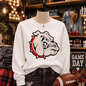 May include: A white long-sleeve sweatshirt featuring a large graphic of a bulldog head wearing a red spiked collar. The bulldog design is rendered in white with black outlines and red accents. The sweatshirt has a crew neckline and long sleeves.