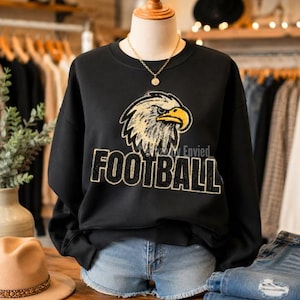 May include: A black long-sleeve sweatshirt featuring a large graphic of a bald eagle head with a yellow beak and the word FOOTBALL in bold, glittery black letters underneath. The sweatshirt is displayed on a mannequin wearing a gold chain necklace.