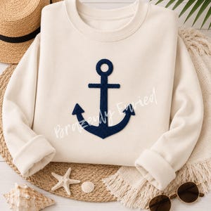 May include: A cream-colored long-sleeve sweatshirt featuring a large, dark navy blue embroidered anchor design centered on the front. The garment has a crew neckline and ribbed cuffs, showcasing a clean, nautical-inspired aesthetic.