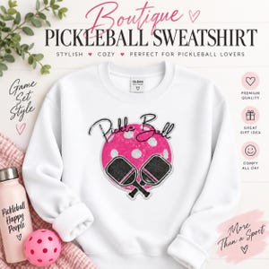 May include: A white crewneck sweatshirt featuring a graphic of a pink pickleball with white dots and two black pickleball paddles crossed in front. The text Pickle Ball is written in a black script font above the graphic on the chest area.