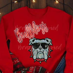 May include: Red sweatshirt featuring a glittery bulldog wearing sunglasses. The word "Bulldogs" is written in a sparkly script above the bulldog graphic. The sweatshirt is a solid red color.
