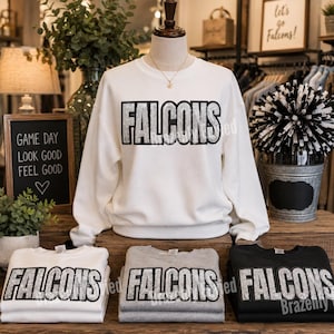 May include: A white long-sleeve crewneck sweatshirt featuring the word FALCONS in large, silver glitter block letters with a black outline. The sweatshirt is displayed on a mannequin, with additional folded sweatshirts in white, gray, and black.
