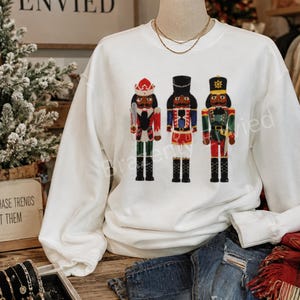 May include: A white long-sleeve sweatshirt featuring a graphic print of three nutcracker figures standing in a row. Each nutcracker wears a distinct uniform in shades of red, green, blue, and gold, complete with tall hats and black boots.