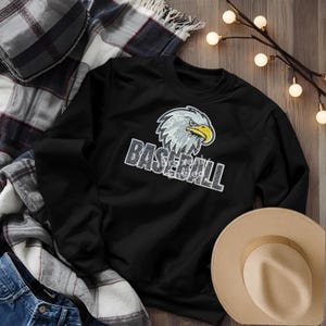 May include: Black sweatshirt with a distressed "BASEBALL" graphic and an eagle head design. The eagle has white and gray feathers with a yellow beak. A tan hat and plaid blanket are in the background.