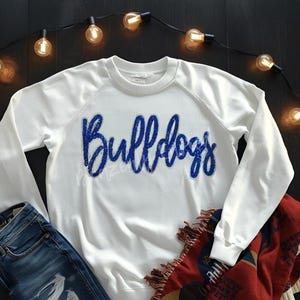 May include: White sweatshirt with the word "Bulldogs" in blue sequined script. The long-sleeved top is paired with blue jeans and a red and cream-colored patterned blanket. A string of lights with warm-toned bulbs is arranged around the items.
