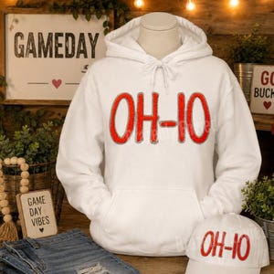May include: A white hooded sweatshirt featuring the red text OH-IO with a grey outline on the chest. The hoodie has a drawstring hood and a front kangaroo pocket. A matching white baseball cap with the same red OH-IO text is also included.