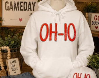 May include: A white hooded sweatshirt featuring the red text OH-IO with a grey outline on the chest. The hoodie has a drawstring hood and a front kangaroo pocket. A matching white baseball cap with the same red OH-IO text is also included.