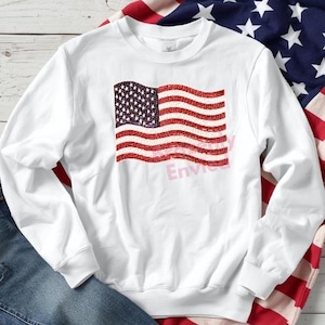 May include: White long-sleeve sweatshirt featuring a graphic of the American flag in red, white, and blue. The flag design is slightly distressed, with the word "Envie" printed in pink across the flag. The sweatshirt is made of a soft material.