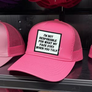 May include: A bright pink trucker hat with a mesh back and a curved brim. The front of the hat features a white square with the text "I'M NOT RESPONSIBLE FOR WHAT MY FACE DOES WHEN YOU TALK" in black.
