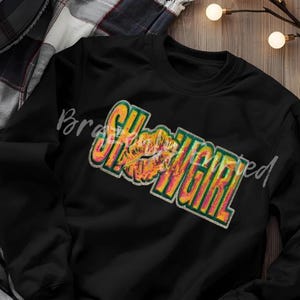 May include: Black sweatshirt with the word "SHOWGIRL" in a colorful, glittery design. The design includes a pair of lips in the center. The sweatshirt is displayed on a wooden surface with string lights.
