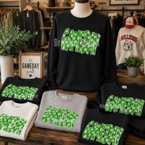 May include: A collection of crewneck sweatshirts in black, gray, and white featuring the word GAMEDAY in green with white polka dots. The text is printed in a bold, blocky font across the chest of each garment, showcasing a consistent design.