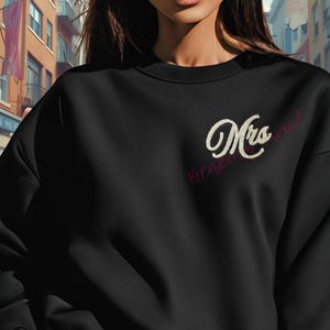 May include: Black sweatshirt with the word "Mrs" in a cursive, rhinestone-like font. The word "Pornzaried" is written in a smaller, cursive, red font below "Mrs". The sweatshirt is a solid black color.
