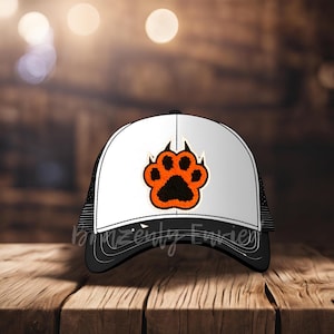 May include: A white and black trucker hat with a black mesh back. The hat has an orange paw print with black claws on the front.