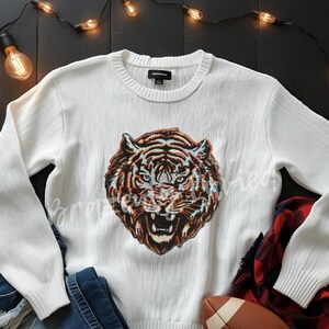 May include: White sweater with a detailed tiger face design in shades of brown, orange, and black. The sweater has a ribbed crew neck and long sleeves. A pair of blue jeans and a football are also in the image.