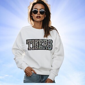 May include: White sweatshirt with the word "TIGERS" in a shimmering, multi-colored, block letter design. The sweatshirt is worn by a person wearing sunglasses and blue jeans.