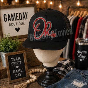 May include: A black baseball cap with a mesh back and a flat brim. The front features an embroidered script logo in red and grey that reads Ohio. The cap is displayed on a neutral-toned mannequin head stand, showcasing the structured crown and adjustable snapback design.
