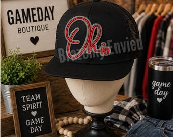 May include: A black baseball cap with a mesh back and a flat brim. The front features an embroidered script logo in red and grey that reads Ohio. The cap is displayed on a neutral-toned mannequin head stand, showcasing the structured crown and adjustable snapback design.