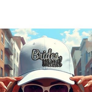 May include: A white baseball cap with the word "Bridesmaid" in a sequined design. The cap is worn by a person wearing sunglasses. The background shows a city street with buildings and a blue sky.