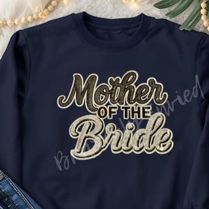 May include: Navy blue sweatshirt with the text "Mother of the Bride" in a decorative font. The text is in a combination of brown and gold colors. The sweatshirt is folded with a pair of blue jeans.
