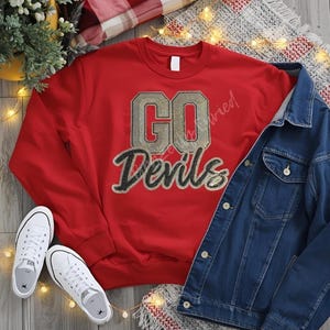 May include: A red sweatshirt with the text "GO Devils" in gold and black lettering, paired with a blue denim jacket and white sneakers. The sweatshirt is the focal point, suggesting a theme of team spirit or casual wear.