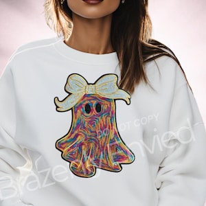 May include: White sweatshirt featuring a colorful ghost design with a bow. The ghost is adorned with a rainbow of colors and has two black eyes. The bow is a light yellow color. The sweatshirt is a casual style.