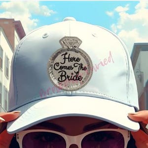 May include: A white baseball cap with a large sequined ring design that says "Here Comes The Bride". The word "married" is written on the side of the cap in pink script. The cap is worn by a person wearing sunglasses.