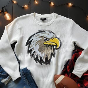 May include: White sweater with a detailed eagle head graphic. The eagle features gray, brown, and yellow accents. The sweater is paired with blue jeans, a red and black plaid shirt, and a football. Decorative string lights are in the background.