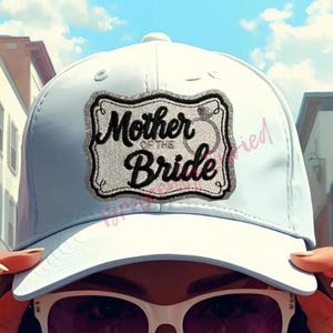 May include: A light blue baseball cap with the text "Mother of the Bride" in a decorative frame. The cap has a curved brim and is worn by a person wearing sunglasses. The background shows buildings and a blue sky.